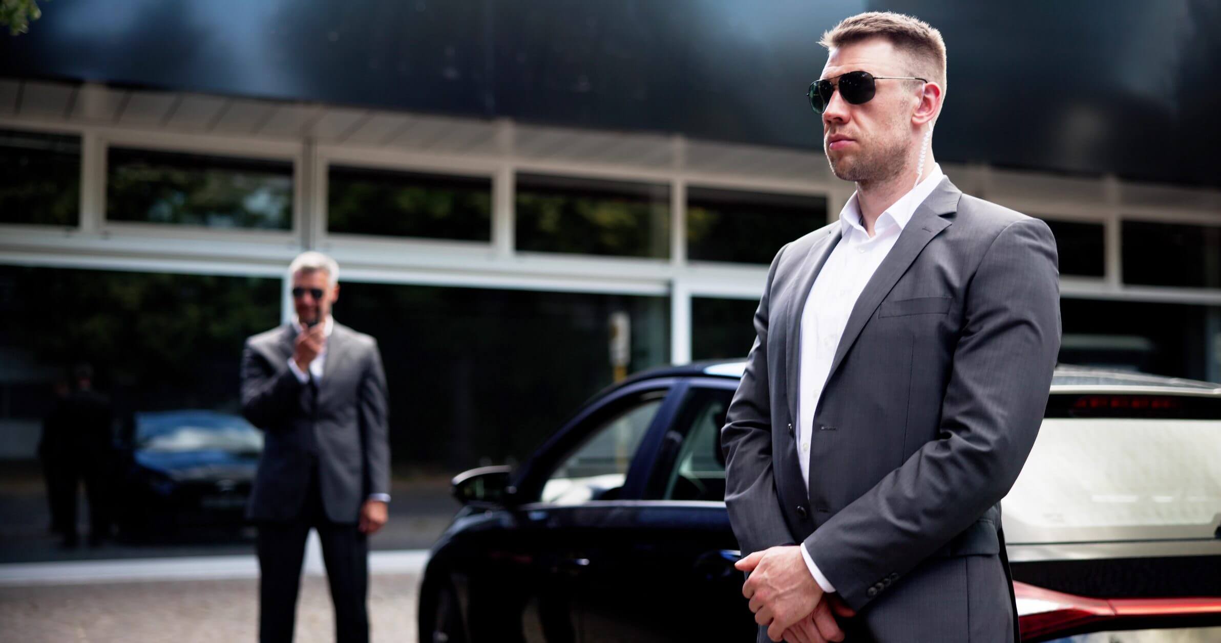 Two security guards in suits near car.