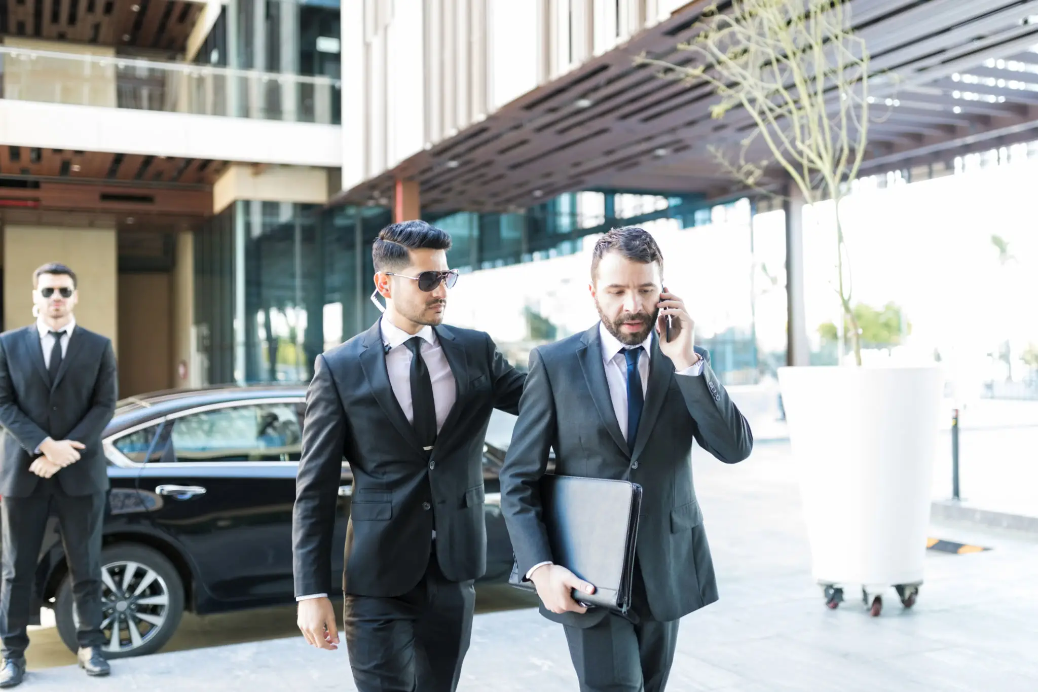 Two businessmen in suits walking with a car in the background.